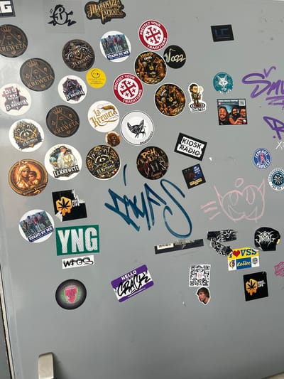 This is a medium-shot, eye-level, full-frame photograph of a collection of stickers on a gray surface, likely a refrigerator door, given the presence of a handle on the right side of the frame. The stickers are varied in shape, size, and design, featuring text, illustrations, and photographs.

Prominent subjects include stylized graphic designs, cartoon characters, and photographs of people. Some stickers have a tattoo or graffiti aesthetic, with bold lettering and intricate patterns. Several circular stickers feature the text "EL KREWETA" and "LA KREWETA" along with imagery of crowns and eyes, suggesting a group or brand affiliation. Another recurring motif is the word "KREWETA GANG".

There are also stickers with text like "KIOSK RADIO," "PROTECT YOUR TRADITIONS," "STAND BY ME," "HHC LIFE," and "YNG." One sticker displays a QR code. A few stickers have a vintage or retro feel, while others appear more modern. The overall impression is that of a personal collection of stickers, possibly accumulated from various sources or representing personal interests and affiliations. The lighting is even, indicating an indoor setting, and there are no obvious weather cues. The image appears to be taken in Brno, Czechia, based on the presence of the word "Brno" on one of the stickers.