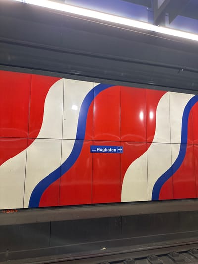 This image captures an unoccupied underground train station platform at Zurich Airport in Kloten, Switzerland. The dominant feature is the wall alongside the tracks, adorned with a repeating graphic composed of large, undulating vertical stripes in red, white, and blue. These stripes are arranged on rectangular panels, creating a flowing, wave-like pattern across the wall.
Centered horizontally on this patterned wall is a blue rectangular sign with white text reading "Zürich Flughafen" (Zurich Airport) and a white airplane icon pointing to the right, indicating direction. The glossy surface of the wall panels reflects the overhead artificial lighting.
Below the wall, the dark grey platform edge is visible, and in the immediate foreground, the train tracks are present, showing sleepers and fastening mechanisms. The overhead ceiling structure is dark grey, with multiple bright, linear fluorescent light fixtures running parallel to the tracks, providing illumination to the scene. No individuals are present on the platform or near the tracks.