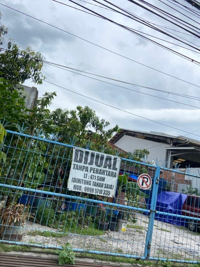 The image shows a blue metal fence in Margahayu, Indonesia, with a sign attached to it that reads "DIJUAL TANPA PERANTARA LT: 471 SHM (DIHITUNG TANAH SAJA) HUB: 0898 5710 335." There is a no parking sign visible through the fence. Green plants are growing on the outside of the fence and behind the fence. The sky is cloudy. The ground is gray and appears to be a concrete or brick patio. There are multiple power lines overhead.