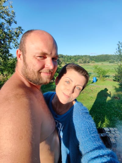 The image is a selfie taken outdoors on a sunny day. A man with a beard, shirtless, and a woman wearing a blue sweater are in the foreground, embracing. They appear to be in a rural setting with green grass, trees, and a blue barrel in the background. There is also a grill with food cooking. The overall mood is relaxed and happy, suggesting a picnic or barbecue.