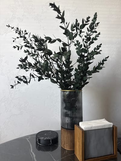In an indoor setting, a dark green, leafy branch arrangement is displayed in a fluted glass vase with a gold rim. The vase rests on a wooden pedestal. To the left of the vase, on a black marble surface, is a stack of round, black marble coasters. To the right of the vase is a wooden napkin holder filled with white napkins. The background is a textured white wall. The scene is devoid of people and activity. The photograph appears to be taken in Velikiye Luki, Russia.