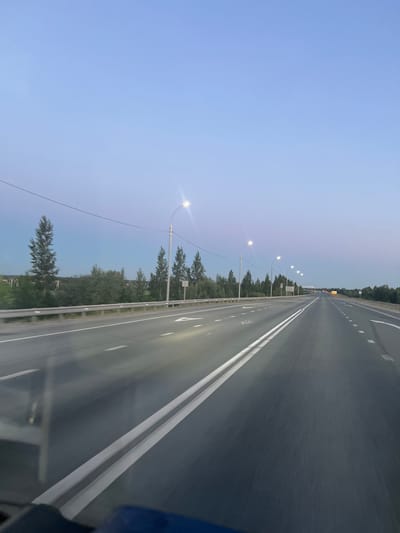 The image shows a view from a vehicle traveling on a highway in Katkovskii, Russia, at dusk. The sky is a soft gradient of blue and pink. Tall, slender pine trees line the left side of the road, interspersed with other greenery. Streetlights with their bulbs illuminated are visible along the highway, with several more stretching into the distance. The highway itself is paved and marked with white lines, indicating multiple lanes. There are no people or other vehicles immediately visible in the frame, but the perspective suggests the road continues into the distance. The overall scene conveys a sense of travel and the quiet ambiance of early evening.
