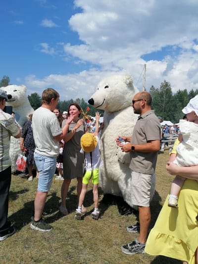 The image depicts an outdoor event, likely a festival, taking place in the city of Krestovskoe, Russia. In the foreground, two individuals in large, furry polar bear costumes are interacting with a group of people. One polar bear appears to be larger and is standing behind a man wearing a grey polo shirt and beige shorts, who is holding a bottle of water. A child in a yellow hat and bright green shorts is reaching up towards the polar bear. To the left, another polar bear costume is visible, partially obscured by a man holding a phone. Several other people, including women and children, are present in the background, with some milling around and others standing near tents or a wooden fence. The ground is covered in dry grass, and a line of trees forms the backdrop against a bright blue sky with scattered white clouds.