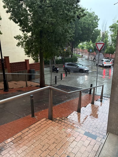 The image depicts a street scene in Esplugues de Llobregat, Spain, on a rainy day. The view is from an elevated position, possibly a building or balcony, looking out onto a street with vehicular and pedestrian traffic.
**Environment:** The street is wet from the rain, reflecting light and creating a sheen on the pavement. Trees line the street, their foliage appearing lush and dark from the moisture.
**Objects:** A grey car is driving through the rain on the street. There's also a white vehicle in the distance. Street signs are visible, including a triangular yield sign. A silver-colored railing runs along the edge of the elevated position from which the photo is taken. A brick-paved sidewalk.
**Location:** Based on the architectural style and street layout, this scene takes place in Esplugues de Llobregat, Spain.
**Action:** Cars are moving through the rain, and it appears that people are walking.