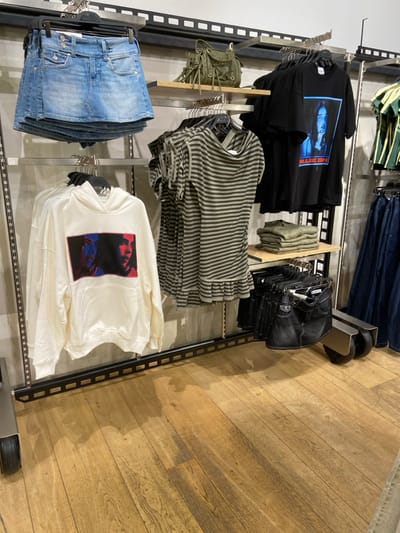 The scene depicts the interior of a clothing store in Almancil, Portugal. The floor is composed of light brown wooden planks. Garments are displayed on metal wall-mounted retail fixtures featuring shelves and hanging rails.

On the upper left, a stack of light blue denim mini-skirts hangs from a multi-tier hanger. To its right, an olive green handbag with fringe details rests on a light wooden shelf. Below this shelf, a series of horizontally striped short-sleeved tops, predominantly olive green and grey, hangs on a rail. Further left and lower, a white hooded sweatshirt is displayed, featuring a central graphic print of two faces rendered with blue and red tones.

On the upper right, multiple black t-shirts hang from a rail, one prominently displaying a blue-toned graphic of Billie Eilish with the artist's name below. Below these t-shirts, a stack of folded olive green clothing items rests on a wooden shelf. In the lower right quadrant, a collection of dark denim skirts or shorts is displayed on hangers attached to a black mobile retail rack, which has visible black wheels. Adjacent to this, a series of dark blue denim trousers or jeans hangs vertically from another rail. A green and white striped short-sleeved top is partially visible on a hanger at the far top-right. No individuals are present in the image.