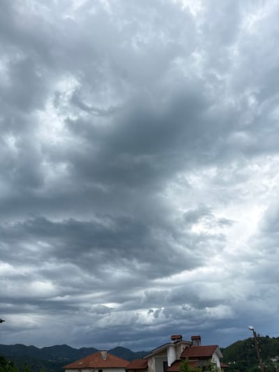 The image shows a cloudy sky over a residential area, presumably in or near Zhaltusha, Bulgaria. The clouds are predominantly gray and layered, suggesting an overcast day. Part of a house is visible on the right side of the image. A utility pole with wires can be seen as well. The roof is tiled with orange/red color. Behind the houses, forested hills rise into the distance.