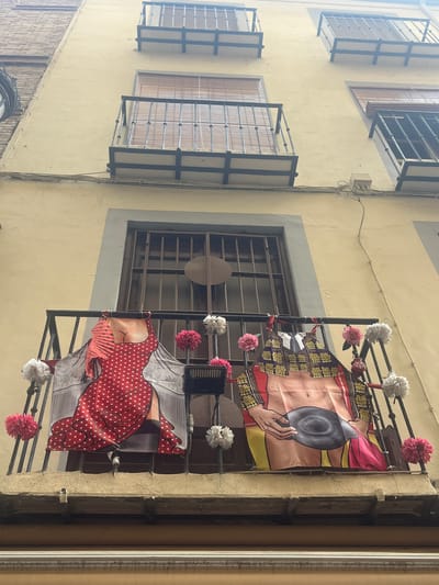 This is a medium-shot, eye-level photograph taken from below, looking up at a balcony on the exterior of a building in the city of Granada, Spain. The building's facade is a pale yellow, with multiple stories featuring balconies with dark metal railings.
On the balcony in the foreground, two large, colorful banners are displayed, hung from the railing. The banner on the left depicts a woman's torso in a red and white polka-dot dress, with a sheer, patterned overlay suggesting a flamenco dress. A red fan is positioned to the left of the banner. The banner on the right displays a bullfighter's costume, with the exposed torso of a man wearing a decorated jacket and holding a black bullfighter's hat. The banner is further adorned with a red cape.
Both banners are decorated with artificial flowers, predominantly in shades of pink and white, attached to the balcony railing. A small black electronic device, possibly a speaker or light, is attached to the railing in front of the woman's banner.
Above the foreground balcony, there is another balcony with blinds visible behind a metal railing, and above that, a third balcony. The building's architecture suggests an older style, with visible brickwork on the left side of the frame and wooden blinds on the windows. Electrical wires run horizontally across the building's facade. The scene appears to be a street or alleyway, given the upward perspective.