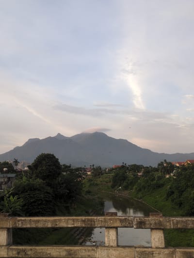 The image depicts a serene landscape in Yên Sơn, Vietnam, featuring a wide river flowing through a lush, green valley. In the foreground, a concrete bridge railing offers a vantage point for the scene. The river, with its calm, reflective surface, meanders towards the horizon, bordered by dense vegetation and trees on its banks. In the mid-ground, a cluster of houses with red-tiled roofs is nestled amidst the foliage, suggesting a small village or settlement. Towering in the background are majestic, mist-shrouded mountains, their silhouettes softened by the ambient light. The sky above is a pale blue, adorned with wispy clouds and a faint sunbeam piercing through, creating a tranquil and picturesque atmosphere. The overall impression is one of natural beauty and rural serenity.