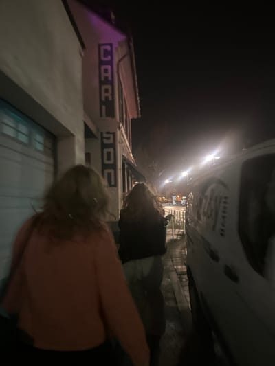 Two women are walking on a sidewalk in front of a building in Bingen am Rhein, Germany. The building has a large sign on the side that says "CARL 500" in block letters. The women are walking in the direction of a white van parked on the sidewalk. There are streetlights illuminating the scene, and the sidewalk appears to be made of concrete. The image is taken at night. It is a dark scene. The women are wearing casual clothing. They are facing forward, walking towards the van. The van is parked on the sidewalk next to a bike rack. The building is a tall, narrow structure. It is made of brick and has a flat roof. The sign is on the side of the building, facing the street. The environment is urban, with sidewalks, streets, and buildings present. The lighting is dim, with streetlights illuminating the scene.