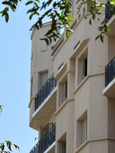 The image is a low-angle, close-up shot of a modern apartment building in Courbevoie, France. The building's exterior is a textured, light beige stucco with geometric architectural details. Several balconies with black metal railings are visible, some with open windows. Green foliage from trees in the foreground partially obscures the building and the clear blue sky. The building appears to be uninhabited.