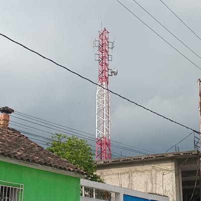 In Duaca, Venezuela, a prominent red and white cellular tower dominates the skyline, equipped with multiple antennas and a parabolic dish. The tower is situated behind a row of buildings, with the foreground featuring a green-painted structure with a tiled roof and a barred window. To the right, another building with a concrete roof and exposed rebar is visible. Overhead, several power lines crisscross the cloudy sky. The overall scene depicts a developed area with infrastructure for communication and residential or commercial structures.