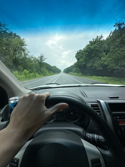 The image shows a first-person perspective from inside a vehicle. A person's hands are visible, one holding the steering wheel and the other resting near a can of a "CARNITINE VITAMIN HIGH PERFOR ENERGY DRINK". The dashboard of the car is visible, including a speedometer with markings up to 240 km/h and a windshield wiper control stalk. Outside the vehicle, a road stretches ahead, lined with lush green trees on either side. The sky is a bright blue with some clouds, suggesting it is daytime and the weather is fair. The perspective, with the road receding into the distance and the trees framing the view, implies the vehicle is in motion on a highway or a similar type of road, likely in a rural or semi-rural setting. The presence of trees and the open road suggest a journey through a natural landscape. The image seems to capture a moment of driving, possibly during a road trip.