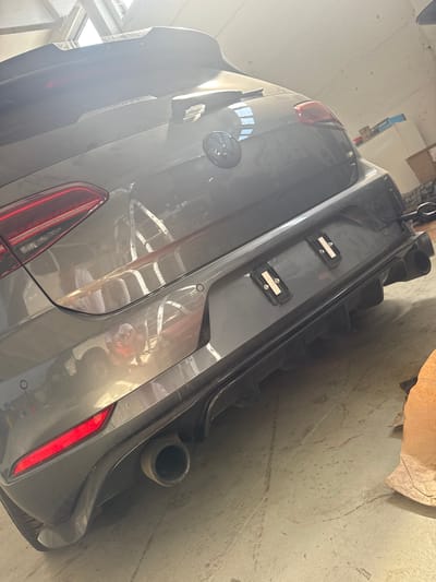 This is a rear-view, medium-shot, eye-level photograph of a grey Volkswagen Golf R hatchback parked indoors. The car is positioned diagonally, facing away from the viewer and towards the right of the frame. The vehicle exhibits a sporty design with a prominent rear spoiler, a black diffuser, and dual exhaust pipes. The license plate area is visible, with two mounting brackets installed, suggesting a license plate may be missing or temporarily removed. The car's taillights are red and illuminated, indicating the brake lights are active or the car is running. The interior of the vehicle appears dusty, with light filtering in from above, possibly from a window or skylight. The car is parked on a concrete floor, which is also dusty and shows some light reflections. The overall environment suggests a garage or workshop. There are no people visible in the image. The scene is in Schkopau, Germany.