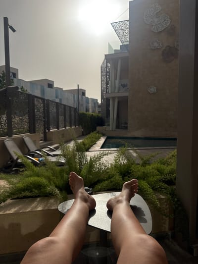 The image depicts a person's legs resting on a gray side table. A thick green fern is visible nearby. In the background, buildings are shown; architectural details of structures, including decorative metalwork and textured walls with organic designs, are present. The image was taken in the city of El Gouna, Egypt. The time of day appears to be daytime, with diffuse sunlight.