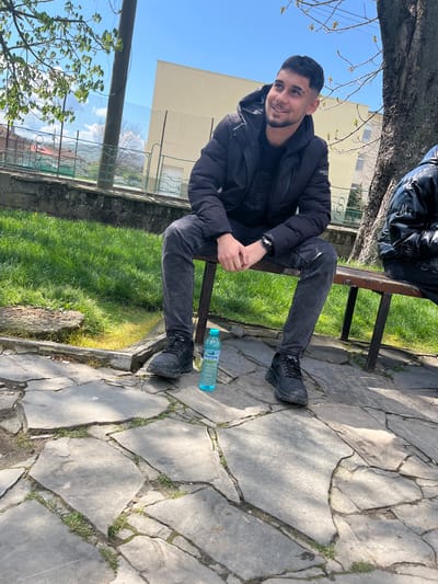 A man sits on a park bench in Dolni Chiflik, Bulgaria. He wears a black jacket and jeans, and black shoes. A water bottle is on the ground next to the bench. The park has green grass, trees, and a stone pathway.