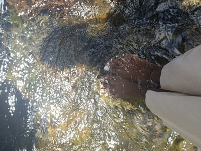 A pair of bare feet, belonging to an individual wearing beige pants, are submerged in a shallow stream of water. The water is clear and flowing, reflecting sunlight, creating a sparkling effect. The streambed is visible beneath the water, composed of rocks and pebbles. The location is a stream within Tsavo North National Park, Kenya, at coordinates -2.984 latitude and 38.023 longitude. The individual appears to be relaxing, enjoying the cool water.