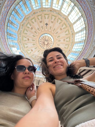 This is a selfie taken from a low angle, looking up at two individuals and a domed ceiling. The subjects are two women, positioned in the foreground and mid-ground of the image. The woman in the mid-ground, facing the camera, has dark hair and is smiling broadly. She is wearing a green, ribbed shirt and a brown woven bag strap across her shoulder. On her left wrist, she wears a dark fitness tracker. The woman in the foreground, wearing sunglasses, has dark hair and is looking upwards. She is wearing a light-colored, ribbed shirt and a white fitness band on her left wrist. Her right hand is resting near her face, touching her hair.
The background is dominated by an ornate, domed ceiling, characteristic of a grand interior space. The ceiling features intricate geometric patterns and floral designs, with a large glass-paned section allowing natural light to filter in. The style of the dome, with its decorative elements and stained glass windows, suggests a historical or architectural landmark. The light indicates it is daytime.
The image appears to be taken indoors, likely in a public building such as a market hall, train station, or museum, given the scale and design of the ceiling. The overall mood is casual and cheerful, with both women smiling. The location context provided, Alboraia / Alboraya, Spain, may hint at specific architectural styles or landmarks within that region. No visible text is present in the image.