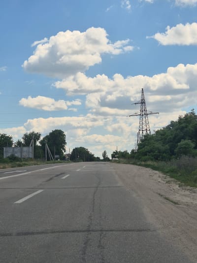 An eye-level, medium-distance shot captures a paved road stretching towards a horizon dominated by a clear blue sky with scattered white clouds. The road, marked with white dashed lines, curves slightly to the right, bordered by a dusty shoulder on the right and grassy verges on the left. In the background, a tall, latticed electrical pylon stands prominently against the sky, surrounded by dense green trees and vegetation. To the left of the road, several concrete poles support overhead wires, and a large, weathered billboard is visible. The scene suggests a rural or semi-urban setting in Ukraine, potentially in or near the city of Pidhaitsi. There are no people or vehicles visible in the frame.