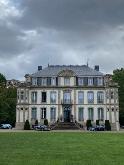 This image depicts the Bugatti Château Saint-Jean, located in Dorlisheim, France. The central subject is a symmetrical, light-colored, three-story stately home or château with a prominent grey mansard roof. The roof features slate tiles, a decorative iron crest along the ridge, two dark chimneys, two finials, and multiple dormer windows. The facade is beige or cream-colored, accented with darker stone banding. A wide stone staircase with ornate railings leads to a central arched entrance door on the ground floor. The second floor exhibits windows with decorative wrought-iron balconies. Octagonal extensions flank the main structure; the left extension includes three circular windows on the ground level. An inscription "1917" is visible above a second-story window to the right of the central balcony.

In the foreground, a well-maintained green lawn occupies the lower portion of the image. Positioned near the base of the central staircase are two dark-colored Bugatti hypercars, resembling Chiron models, parked facing generally towards the viewer. To the far left, on a paved surface, a light blue or silver Porsche 911 GT3 (992 generation) is partially visible. Four dark green, cone-shaped topiary shrubs are evenly spaced in front of the building's lower facade, two on each side of the stairs. Dense green foliage from trees borders the property on both the left and right sides. The sky is entirely overcast with a uniform grey cloud cover. No individuals are present in the scene.