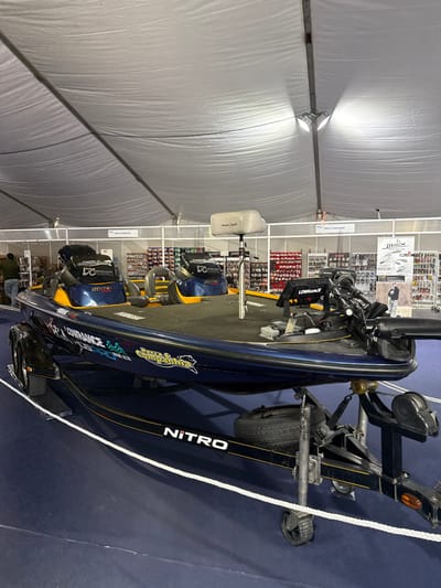 The image depicts an indoor exhibition space, identified as a fishing event in Mora, Portugal, held within a large white event tent illuminated by overhead industrial lights. The central subject is a dark blue and yellow Nitro bass boat prominently displayed on a black dual-axle Nitro trailer. The boat is equipped with multiple fishing seats, including an elevated pedestal seat, a trolling motor at the bow, and Lowrance brand electronic navigation/fish-finding equipment at the helm. Decals on the boat's hull include "YAMAHA," "LOWRANCE," "SIMRANCE," "DC," "CHAMPIONSHIP," and "PESCA & COMPANHIA." The trailer features a horizontally mounted spare tire. A white rope barrier delineates the boat's display area on the blue carpeted floor. In the background, several retail display racks are visible, stocked with a variety of fishing lures, tackle, and accessories. Signage in the background includes "PESCA E COMPANHIA," "molix," and "MEGLEAN." A single individual, partially obscured, stands among the background displays on the far left. No direct actions or interactions are occurring.