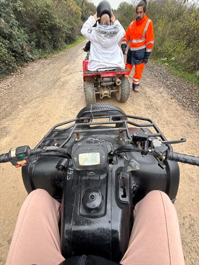 An overhead, first-person view depicts a line of all-terrain vehicles (ATVs) on a dirt path in Çiftlikköy, Turkey. In the foreground, the lower torso and legs, clad in light brown sweatpants, are visible from a seated position on a black ATV. The handlebars, control panel with a display, and a front cargo rack with a secured spare tire are visible on this ATV. Directly ahead, a red ATV is positioned, facing away. A person wearing a white winter jacket with a fur-lined hood is seated on the red ATV, hands raised near their head, seemingly adjusting a dark hood or helmet. The red ATV bears the alphanumeric identifier "LABR 041" on a rear plate. To the right of the red ATV, a male individual stands on the dirt path. He wears a bright orange high-visibility jacket and trousers with dark blue accents, and work boots. He has dark hair tied in a bun and a beard, and is looking towards the person on the red ATV. The path is unpaved, appearing damp and muddy, bordered on both sides by dense green foliage and trees under an overcast sky.