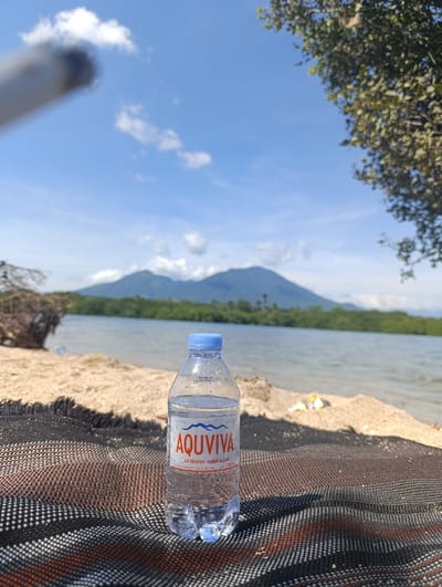 The image shows a bottle of "AQUVIVA" air mineral murni alam water on a black and brown striped picnic blanket. The bottle is centered in the image and is sitting on the blanket, which is spread out on a sandy beach. The background shows a body of water, likely a lake or the ocean, with a line of trees and a mountain range in the distance. The sky is blue with white clouds. The location is a beach in Kecamatan Banyuputih, Indonesia. The beach is covered in sand with some vegetation and small pieces of litter scattered across the sand. The sun is shining brightly. The overall atmosphere is serene and peaceful. The scene is consistent with a relaxing outdoor environment. No people are present. A blurry object appears to be a cigarette in the upper left hand corner of the image.