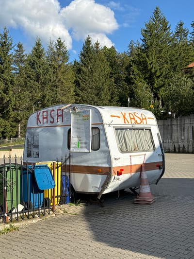 The image depicts an outdoor scene in Karpacz, Poland, featuring a weathered white and brown camper trailer, presumably functioning as a ticket or cash office. The trailer has "KASA" painted in red on its front-left side and in orange on its rear-right side. A white sign labeled "REGULAMIN" (regulations) is affixed to the front-left side of the trailer. The rear of the trailer displays a window with closed, light-colored curtains and is supported by what appear to be blocks or stands, resting on a paved surface of grey interlocking bricks. A worn orange and white traffic cone, possibly stacked, is positioned behind the trailer. To the left foreground, partially obscured by a black metal fence, are three wheeled garbage bins: one green, one blue, and one yellow. The background is dominated by a dense stand of tall evergreen trees, consistent with a mountainous environment. To the right, a grey concrete wall with "BUS" repeatedly stenciled in white is visible. The sky is blue with scattered white cumulus clouds, and the scene is brightly lit by sunlight, casting shadows. No individuals are present in the image.
