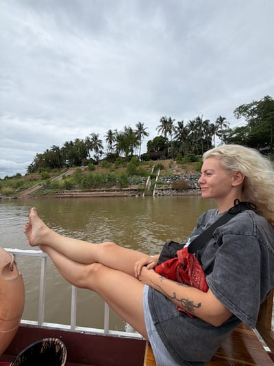 A blonde woman with windswept hair relaxes on a boat on a murky river, her legs crossed and propped up on a white railing. She is wearing a grey t-shirt and denim shorts, with a red bandana peeking out from a black bag slung across her body. The boat is moving, evidenced by the water's ripples. In the background, a lush green hillside is covered in palm trees and dense vegetation, with a few buildings nestled amongst them. A concrete staircase leads up the hill, suggesting the location is a riverside village or town. The sky is overcast, casting a soft, diffused light over the scene. The boat appears to be navigating a section of the Mekong River near Luang Prabang, Laos, a region known for its scenic beauty and tranquil atmosphere.