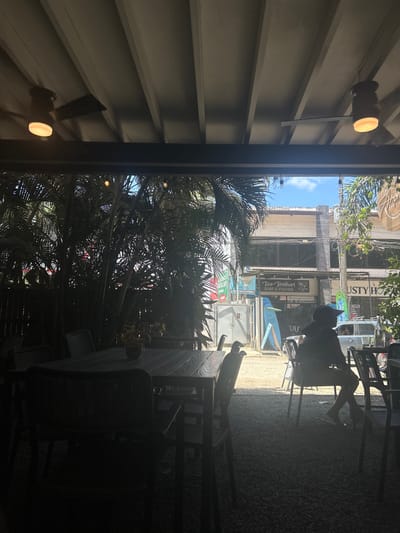 The image shows a patio with a black wooden table and chairs. The table is set with glasses and plates. A person is sitting in a chair on the patio, facing away from the camera. The patio is located under a covered structure with a ceiling fan. The image is taken from a point of view looking out from the covered area. The patio is situated in front of a building with the words "Taco Brothers Surf & Fishing" visible on the facade. Green foliage is seen behind the building, filling the background. The location is in the Cobano District of Costa Rica.