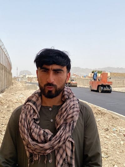 A medium shot shows a man in a dark green, button-down shirt with a brown and white patterned scarf. He is standing near a newly paved road. In the background, there are two road rollers and a man in blue clothing standing on one of the rollers. A concrete wall with barbed wire runs along the edge of the road. In the distance, there are buildings and mountains under a hazy sky. The location is قندهار, Afghanistan.