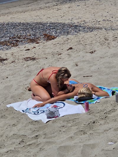 Two women are on a sandy beach in San Clemente, California, United States of America. The woman in the foreground is lying on her back on a striped towel, wearing a blue bikini top and bottom. She has blonde hair and a tattoo on her back. The woman behind her is kneeling and leaning over her, wearing a red bikini. Her hair is also blonde. She appears to be touching or massaging the other woman's back. A white towel with a graphic design is spread out on the sand, and a gray folded item and a pink can are on top of it. Another pink can is placed on the striped towel. The beach is covered in sand and small pebbles, with some dried seaweed scattered around. The ocean is visible in the upper left corner of the image.