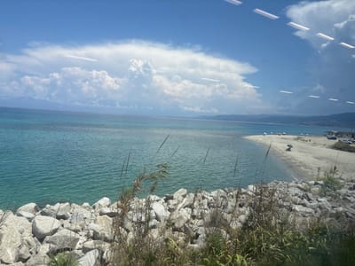 The image depicts a serene coastal scene in Briatico, Italy, bathed in the bright light of a sunny day. The sea, a vibrant turquoise, gently laps against a rocky shoreline in the foreground. Beyond the rocks, a sandy beach curves into the distance, dotted with colorful umbrellas and a few beachgoers enjoying the leisurely atmosphere. The water is calm, reflecting the clear blue sky overhead, which is adorned with fluffy white clouds. In the far distance, rolling hills, possibly part of the Italian coastline, rise from the horizon. The overall mood is peaceful and idyllic, evoking a sense of summer vacation and relaxation. The time of day appears to be late morning or early afternoon, given the strong sunlight. There are no visible text elements in the image.