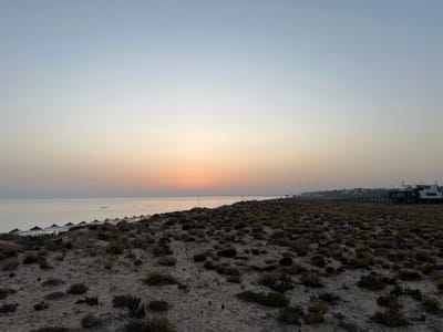 This image depicts a coastal landscape at either sunrise or sunset, specifically in the area of Almancil, Portugal. The foreground is comprised of sandy dunes sparsely covered with low-lying, dry, dark green and brown shrub vegetation. To the left, along the shoreline, a series of uniformly spaced white beach umbrellas or sunshades are lined up near the calm, reflective grey-blue water of the Atlantic Ocean. A small, light-colored boat is visible on the water further offshore.

In the mid-ground on the right, a dark-colored wooden boardwalk extends parallel to the coastline, leading to a complex of modern, multi-story buildings with dark exteriors and flat roofs, possibly commercial establishments like beach clubs or restaurants. One large building features multiple levels and a prominent outdoor deck area. Distant, lighter-colored structures are faintly visible on the coastline in the far right background. The sky exhibits a clear gradient, transitioning from a light blue at the zenith to a pale orange and pink hue directly above the horizon, where the sun's light source is implied but the sun itself is not directly visible. No individuals or specific actions are discernible within the frame.