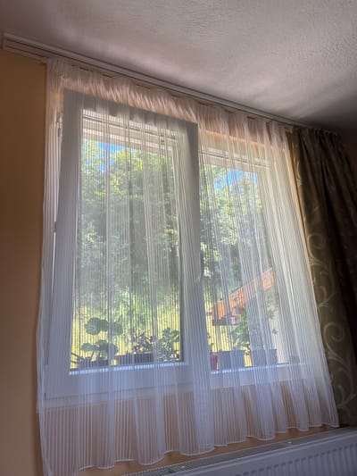 The image shows a window with sheer white blinds in what appears to be a residential setting. Through the blinds, glimpses of greenery, possibly trees and vegetation, can be seen, along with a glimpse of a red roof. The light is bright, suggesting it is daytime. The window is located in a room with beige colored walls. Above the window is a decorative curtain. To the right of the window there is a white radiator. While the photo was taken in Aldomirovtsi, Bulgaria, there are no explicit details in the image that verify this location.