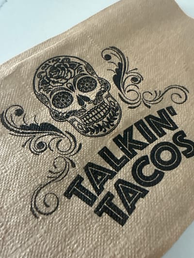 The image shows a tan-colored napkin with a sugar skull design and the words "TALKIN' TACOS" printed in black ink. Ornate black swirls also decorate the napkin. The exact location within Davie, United States of America is not discernable from the image.
