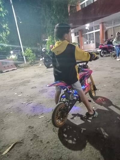 This image captures a nighttime scene in a paved outdoor area adjacent to a building in Nganjuk, Indonesia. The primary subject is a child, viewed from the rear-right, seated astride a stationary pink and black mini dirt bike with gold-colored wheels. The child is wearing a yellow and black hooded jacket, light-colored shorts, and patterned open-toed sandals, with their feet flat on the ground to support the bike. Blue LED lights are visible emanating from beneath the bike's chassis, casting a blue glow on the pavement.
In the background to the left, a tall white street light pole extends upwards, surrounded by dense green foliage. A reddish-brown plastic barrier rests on the ground near the left frame edge. To the right of the child, a multi-story building with a reddish-brown facade and numerous windows is visible. Several motorcycles are parked in front of this building, including a prominent red scooter. Further back on the right, an adult female in a light-colored hooded top and blue jeans stands near a smaller wheeled vehicle, possibly another mini-bike. The ground is a dark, uneven paved surface with minor debris. The scene is illuminated by artificial light sources, casting distinct shadows, indicative of nighttime conditions.
