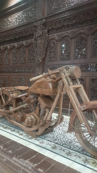 This image depicts an indoor scene in Glantengan, Indonesia. The primary subject is a meticulously crafted, full-scale wooden motorcycle sculpture, rendered in a light brown wood. The motorcycle features detailed representations of a fuel tank, engine, handlebars, headlight, and spoked wheels; the word "HARLEY" is faintly discernible on the fuel tank. The sculpture rests on a patterned tiled floor, which consists of repeating grey and white geometric and floral motifs, bordered by a strip of off-white tiles with dark scrollwork. In the immediate foreground, a low step or barrier constructed of distressed, reddish-brown tiles is visible. The background is dominated by a wall composed of dark brown, intricately carved wooden panels, displaying elaborate floral and abstract designs, including arched elements, consistent with traditional Indonesian woodwork. No individuals or active interactions are present in the scene.