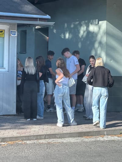 A group of about eight teenagers stands along a building in Liptovský Hrádok, Slovakia. They are positioned on a paved sidewalk next to the building and an adjacent asphalt street. The group appears to be standing close together. They are dressed in contemporary casual clothing, including jeans, shorts, T-shirts, and a jacket. The building is a light gray color with a metal door and a sign with the number "895" on it. The lighting suggests it is daytime.