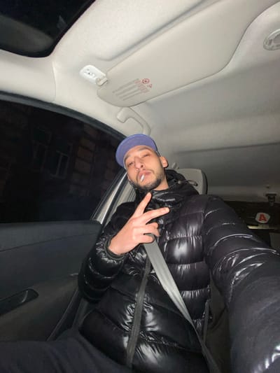 A man with a beard and a blue baseball cap, seated in a car, wears a glossy black puffer jacket and a grey seatbelt. He is holding up a peace sign, and there appears to be something in his mouth, possibly a cigarette. The interior of the car includes a white sun visor and a black car door. Nighttime is visible through the car window.