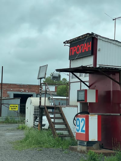 The image shows an outdoor propane gas station in Kopeysk, Russia, under a cloudy sky. A prominent red LED sign on top of a building displays the word "ПРОПАН" (Propane) in Russian. A gas pump with a "92" label is visible to the right, along with a smaller digital display on the side of the building. To the left, a yellow sign reads "ПРИЁМ ЛОМА" (Scrap Metal Acceptance) and points towards a building. A white car is parked near a structure with barred windows. The area appears to be somewhat industrial, with various metal structures and overgrown grass.