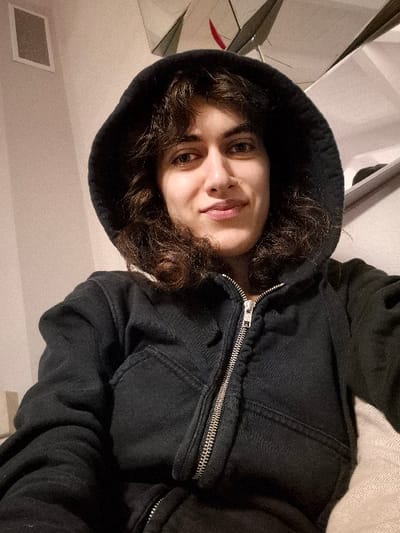 A single individual, an adult female with medium-dark, curly hair, is depicted in a close-up, selfie-style indoor photograph taken in Châtelaillon-Plage, France. She wears a black hooded sweatshirt with the hood pulled over her head, partially covering her hair. The sweatshirt features a partially unzipped silver-colored zipper. The subject faces the camera directly, maintaining eye contact and exhibiting a faint smile. The background on the left side shows a light-colored wall with a white rectangular ventilation grille or speaker mounted high. To the upper right, an angled reflective surface, possibly a mirror, reflects indistinct white and dark shapes. A light-colored, textured surface, likely upholstery, is visible in the lower right, suggesting the subject is seated.