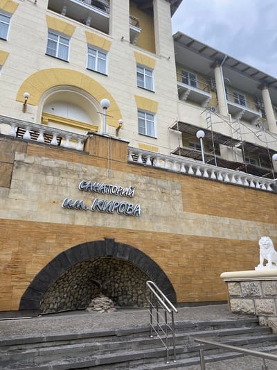 The image shows a building with a sign that reads "САНАТОРИЙ им. КИРОВА" (Sanatorium named after Kirov) in large, silver, three-dimensional letters. The building's facade is light yellow with yellow arched decorative elements above windows. Below the sign is a stone-clad arched alcove containing a statue of an eagle perched on rocks. To the right of the alcove, a white lion statue sits atop a stone pedestal. Stone stairs with metal railings lead up to the entrance area. The background shows a cloudy sky. The building is located in the city of Zheleznovodsk, Russia.