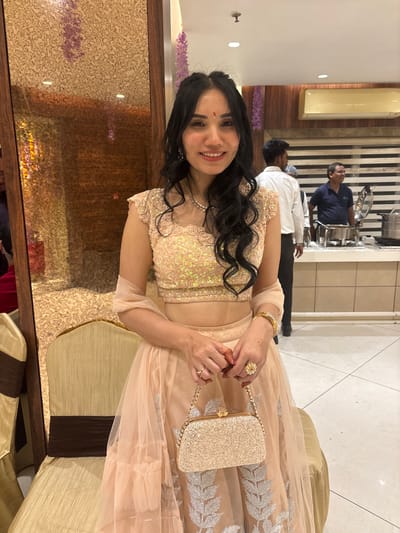 A woman smiles, adorned in a peach-colored lengha with a sequined bodice. She holds a sparkly gold purse by its chain handle. Her hair is black and wavy, and she wears jewelry: a necklace, earrings, a bracelet, and two rings. Two men in casual clothing are visible in the background, near a buffet setup with warming trays. The photo is taken at a slight angle, seemingly in a restaurant or event venue.