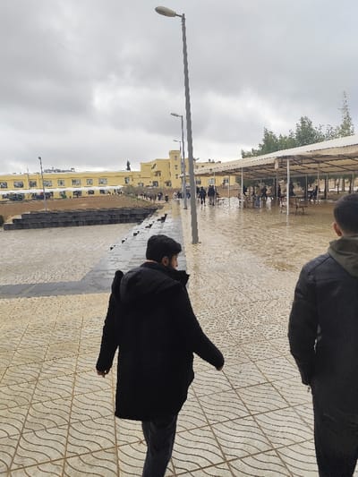 The image depicts an outdoor environment in Jibb as-Safa, Syria, under an overcast sky. The ground in the foreground is a light-colored paved pathway with a wavy, patterned texture, exhibiting wet areas consistent with recent precipitation.

Two individuals are prominent in the immediate foreground, observed from behind as they walk away from the camera. The person on the left wears a black long coat and dark trousers; their dark hair and beard are visible as their head is slightly turned. The person on the far right is partially visible, wearing a dark jacket over a dark hooded garment.

Extending into the mid-ground is a broad paved walkway. Parallel to this walkway, multiple grey streetlights with overhead fixtures are erected. To the left of the main pathway, a dark-colored, multi-tiered structure resembling stepped seating or planters is visible. Further along the pathway, a small, dark-colored dog is observed walking. To the right of the walkway, a large, open-sided structure with a white fabric canopy roof and white support poles covers a paved area furnished with tables and chairs, under which several individuals are sitting and standing.

In the mid-ground and background, a distinctive yellow multi-story building with numerous windows dominates the horizon. A dark statue or monument stands on the ground in front of this building. Additional yellow buildings are partially visible to the right. Multiple individuals are distributed across the paved areas and pathways, engaged in walking, standing, and congregating near the buildings and under the covered structure. Green trees are visible behind the covered structure and in the distant background.