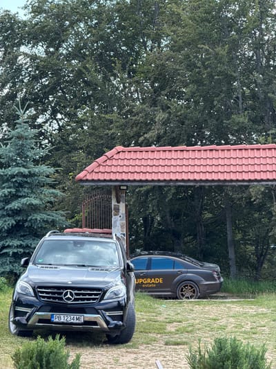 In Bukovo, Bulgaria, a black Mercedes-Benz ML-Class SUV with license plate PB 1234 ME is parked on a grassy area in front of a gated entrance to a property. Behind the SUV, a dark-colored sedan with "UPGRADE estate" emblazoned on its side is parked further into the property. The entrance is covered by a red-tiled roof and a metal gate. The background is dominated by lush green trees, suggesting a rural or semi-rural setting. A blue spruce tree stands to the left of the SUV. The sky is overcast.