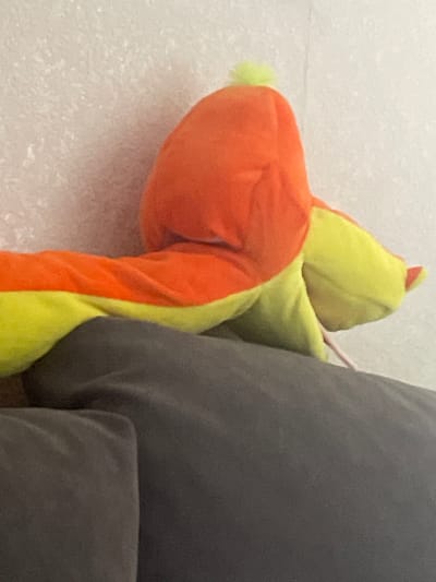 The image shows a close-up view of a stuffed toy, primarily orange and yellow, resting on top of a dark gray surface, possibly a sofa or cushion. The toy has a round, bulbous head with a small, bright yellow tuft on top, and an elongated body with alternating orange and yellow sections. The background is a textured, light-colored wall. There is no indication of the city of Ардонь, Russia, or any urban environment in the image.