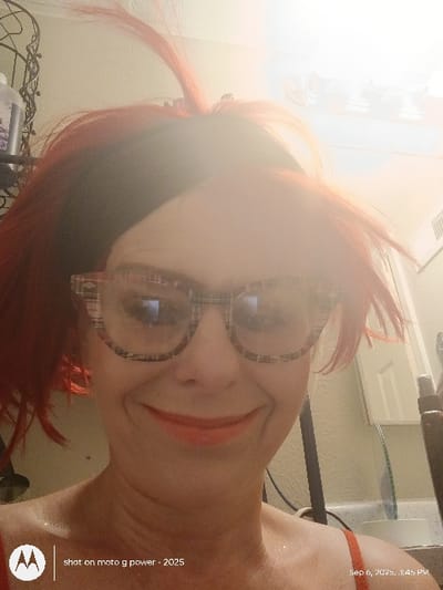 This image is a close-up selfie of an adult female located in Westminster, United States, taken indoors. The subject has bright red hair, appearing disheveled with strands extending upwards, and a visible dark band at the crown. She wears large, thick-framed glasses with a light plaid pattern, which reflect an overhead light source. Her face shows a broad, closed-lip smile with orange-red lipstick. A red strap, consistent with a camisole or bra, is visible on her right shoulder. The background features a light-colored wall with various objects. To the left, a dark, possibly metallic, decorative item and a bottle are visible. To the right, a white ventilation grate or light fixture is present above a light-colored cabinet or door. Below this, a dark cylindrical object stands near a visible counter edge and what appears to be a light-colored sink or basin. The overall lighting is bright, causing significant glare across the top of the subject's head and face. A watermark "shot on moto g power - 2025" is present in the lower left, and "Sep 6, 2025, 3:45 PM" is in the lower right.