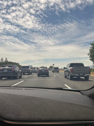 The image is a view from the inside of a vehicle traveling on a highway. It shows a traffic jam with multiple cars and trucks in front. The sky is blue with scattered white clouds. The highway appears to be in a rural or suburban area, with some vegetation and trees visible on the side. The time of day appears to be daytime, likely midday, given the lighting conditions.
