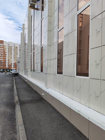 A low-angle, eye-level shot shows the side of a modern building with a light-colored tiled facade featuring a marbled pattern. Tall, narrow windows with reflective, amber-tinted glass are interspersed across the facade. The building appears to be commercial or residential, with a few residential buildings visible in the background. To the left of the building is a paved walkway and a street. A woman is walking away from the camera on the sidewalk, carrying a bag. A white van is parked on the street. The sky is cloudy, with some patches of blue. The scene suggests an outdoor setting in a developed urban area, likely during daylight hours.