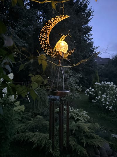 A solar-powered moon-shaped wind chime with a glowing bulb is hanging from a tree branch in a garden at dusk. The wind chime is made of dark metal with intricate cutouts. The garden is lush with dark green foliage, including bushes and trees. In the background, there is a house with a dark roof and windows visible, suggesting a suburban or rural setting. The sky is a deep blue, indicating twilight. The scene conveys a peaceful and serene atmosphere. The location is identified as Romashkino, Russia.